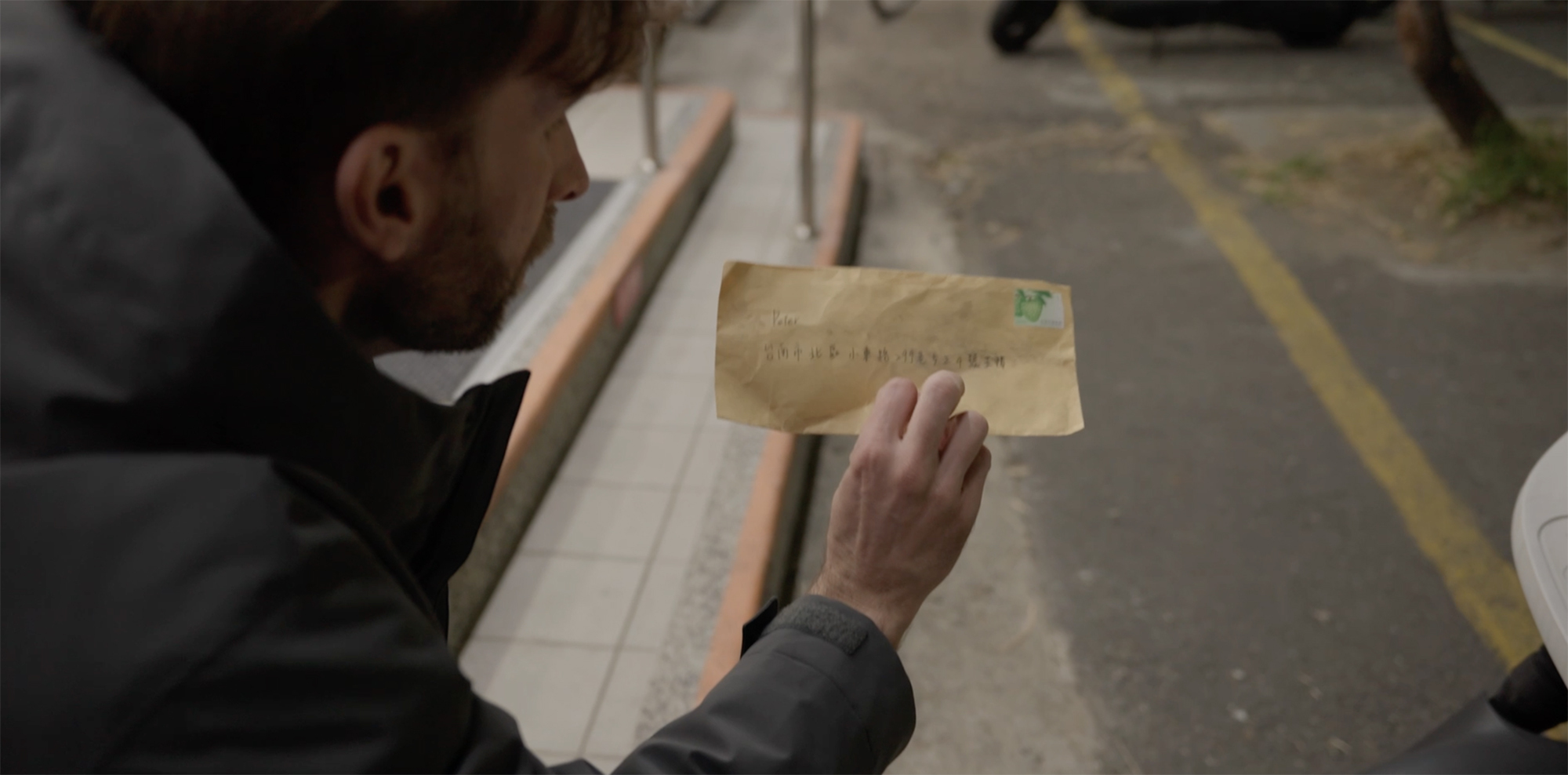 Person in grey jacket holding brown envelope whilst examining white papers on desk surface.