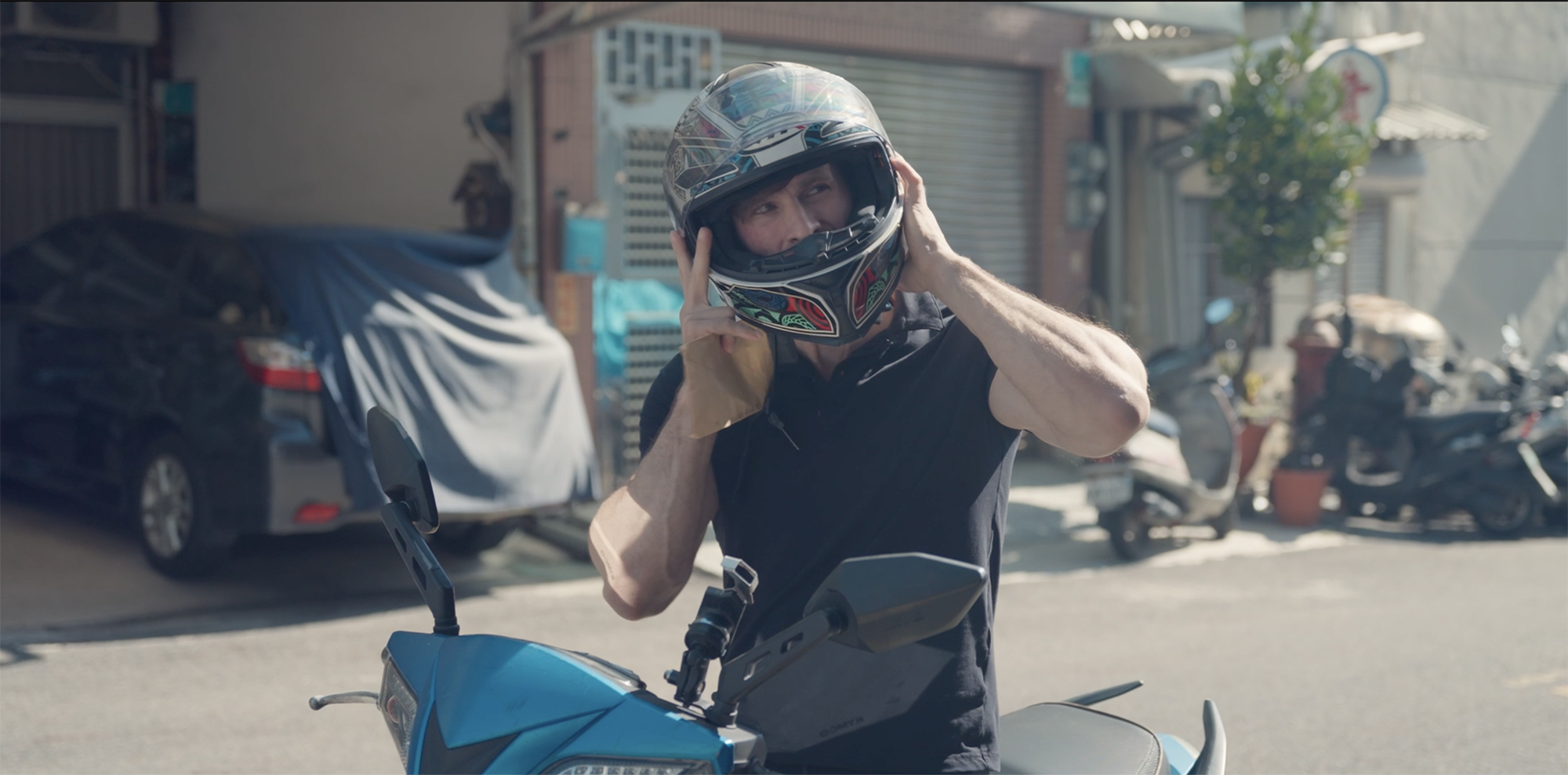 Man on blue motorbike putting on helmet, sunny street scene with shops and parked vehicles in background.