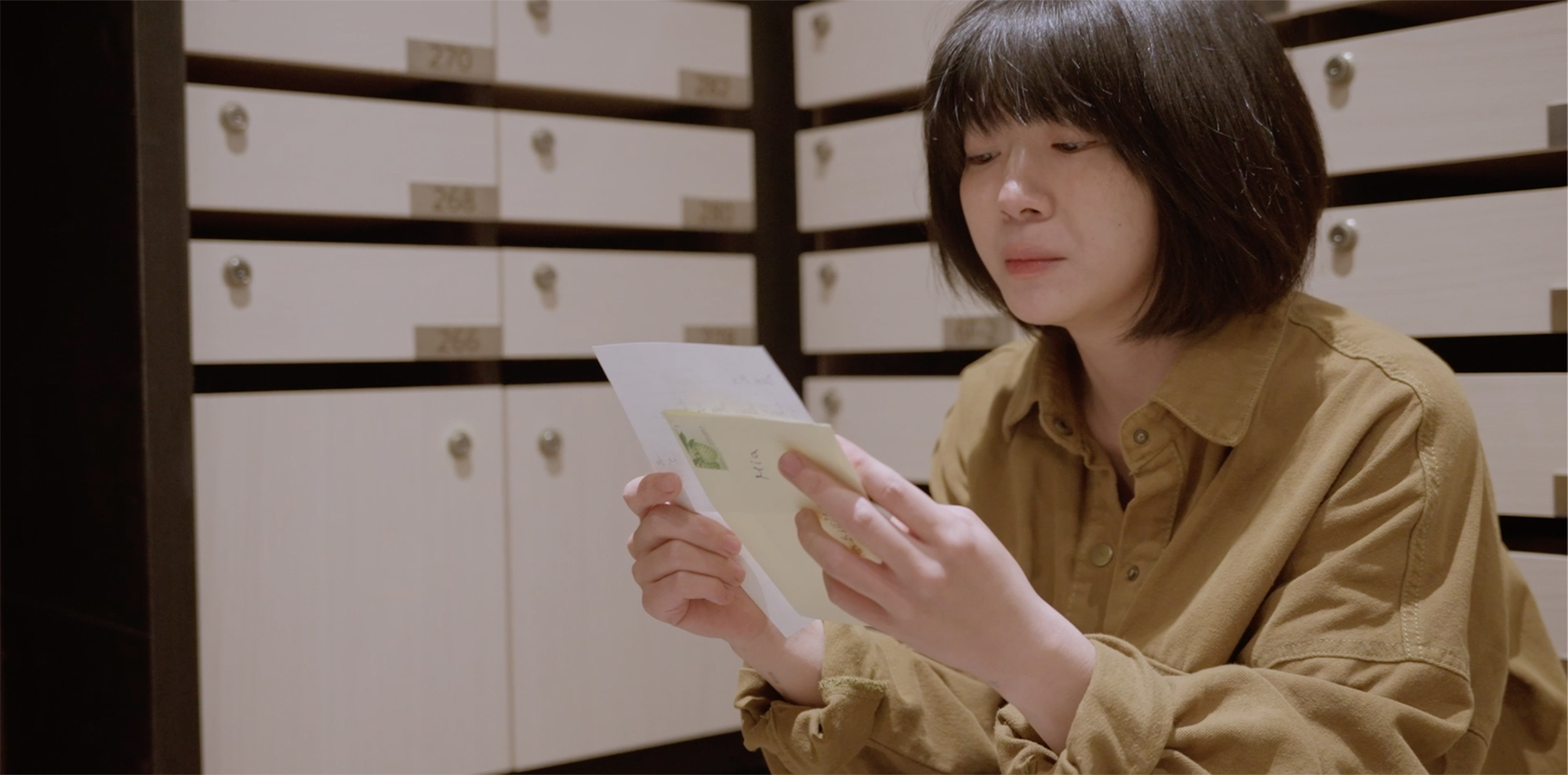 Woman with dark bob haircut wearing mustard yellow shirt reading white paper whilst sitting beside white filing drawers with dark handles.