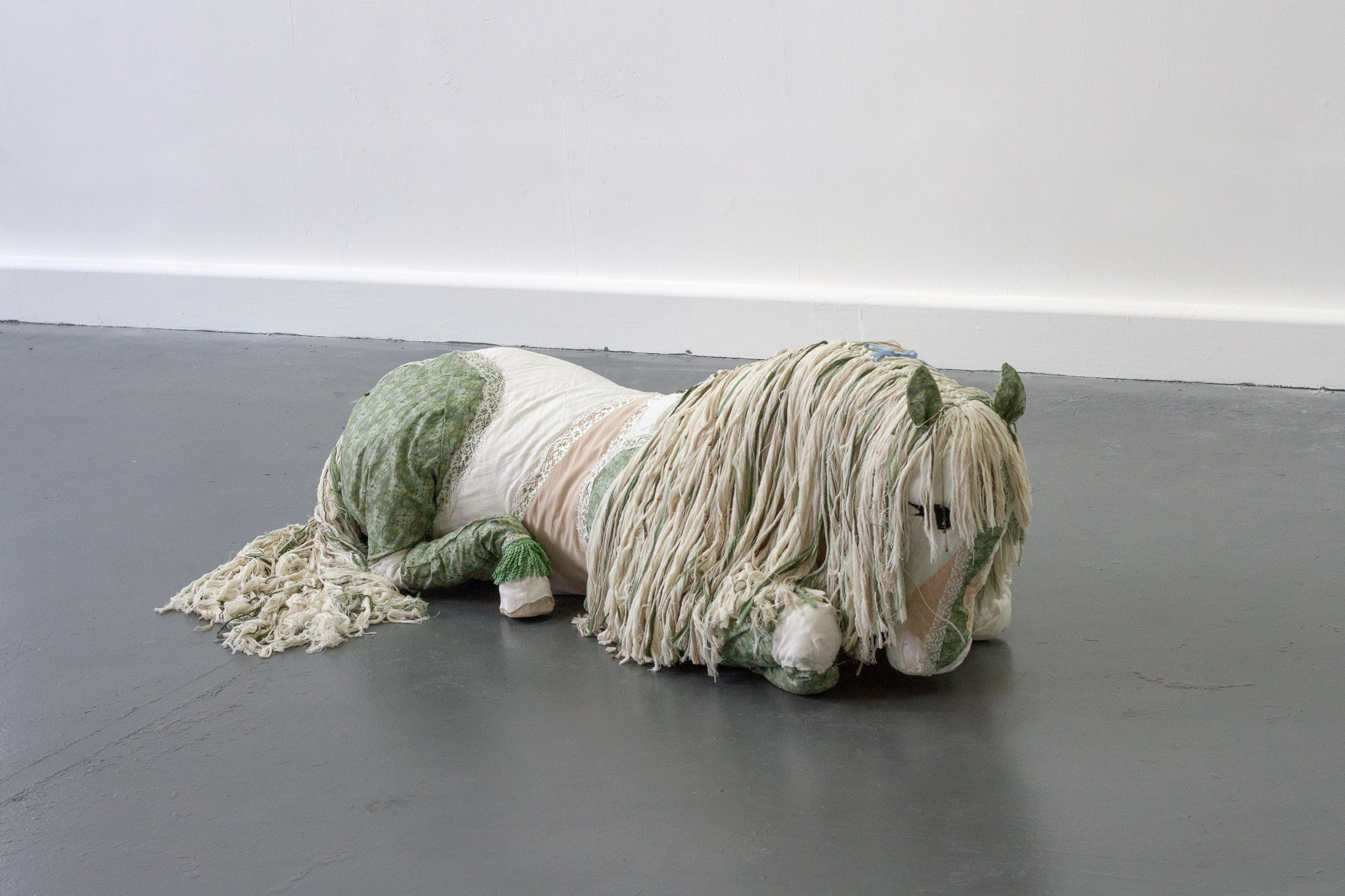 Sculpture of a horse made from soft materials lying on a grey gallery floor.