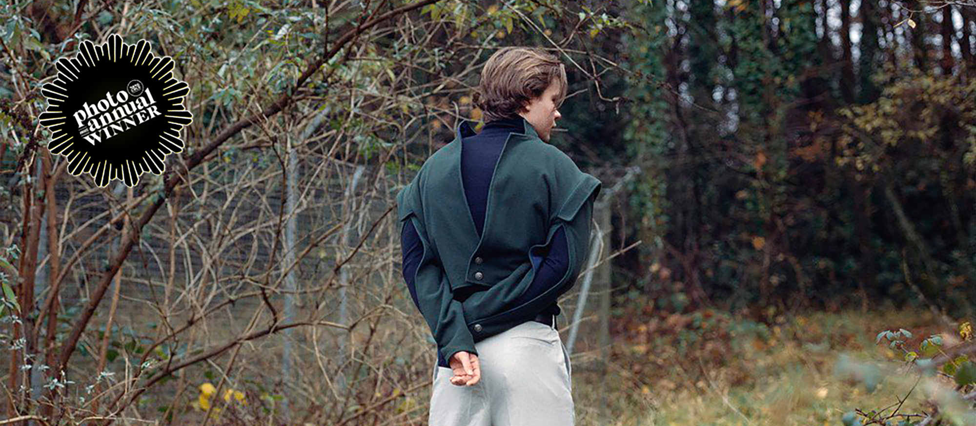 A photo of a person with their back to the camera and their arms behind their backs standing in a woodland. A logo with text 'Photo annual winner' appears in the top-left corner.