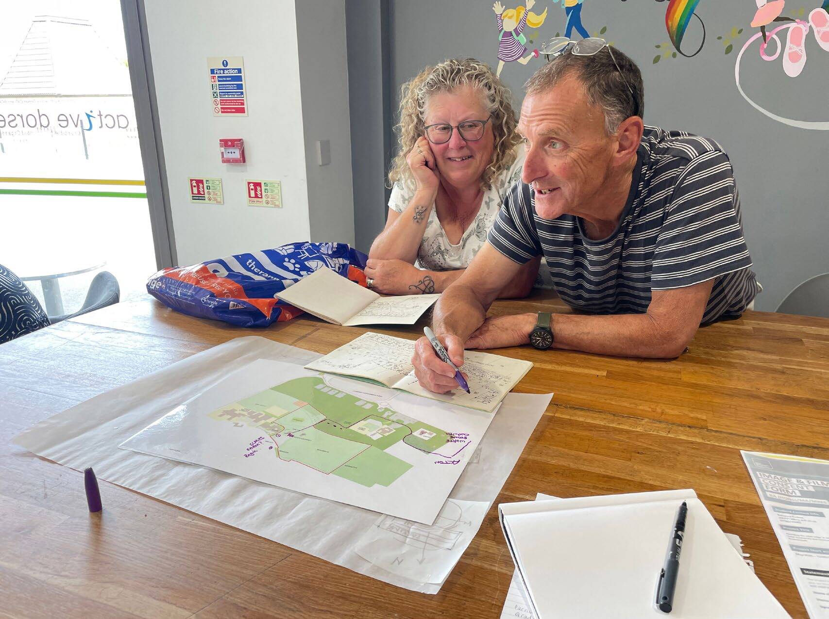 Middle-aged couple examining green architectural plans on wooden table in bright office with colourful wall decorations.