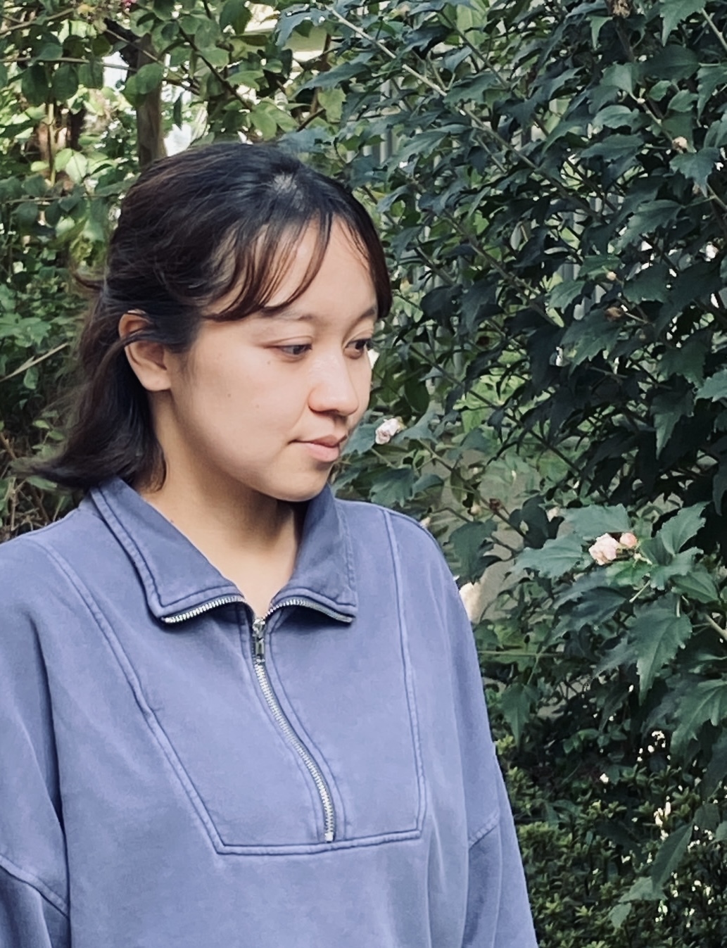 Woman in blue zip-up top with dark hair looking to the side, standing in front of dense green foliage.