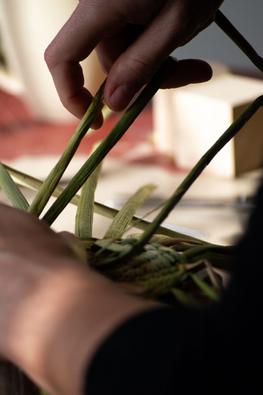 Hands weaving green palm fronds in a basket, with blurred background of people and pink fabric.