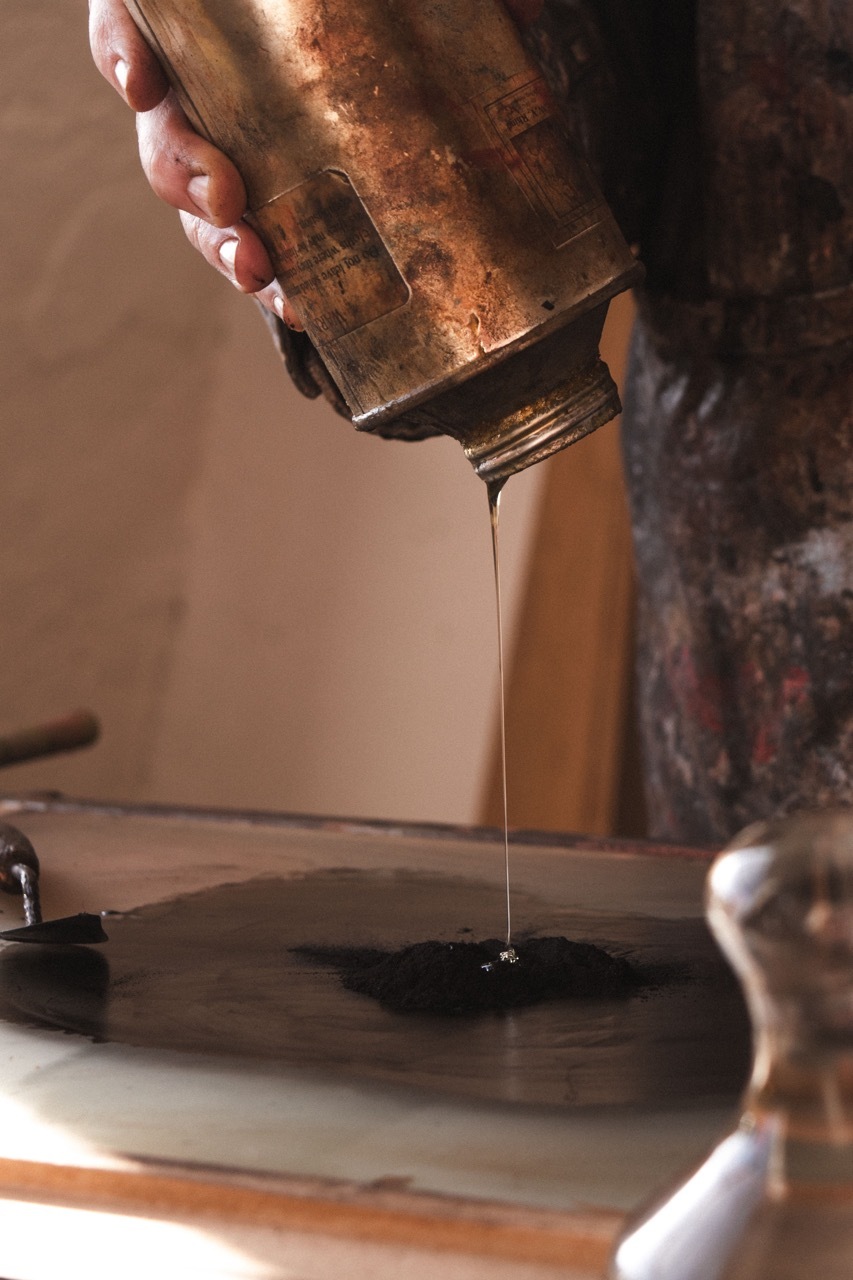 Hands holding weathered copper vessel pouring thin stream of dark liquid onto flat surface in warm-lit workshop setting.