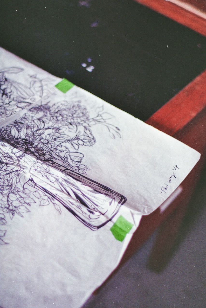 Sketchbook open showing detailed pen drawings of trees and foliage with green tape securing corners to dark surface.