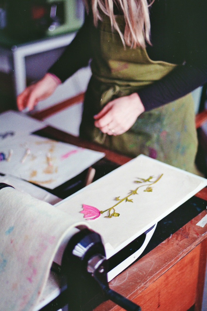 Person in olive green jumper sitting at desk with scattered papers, one showing pink flower illustration, blurred background.