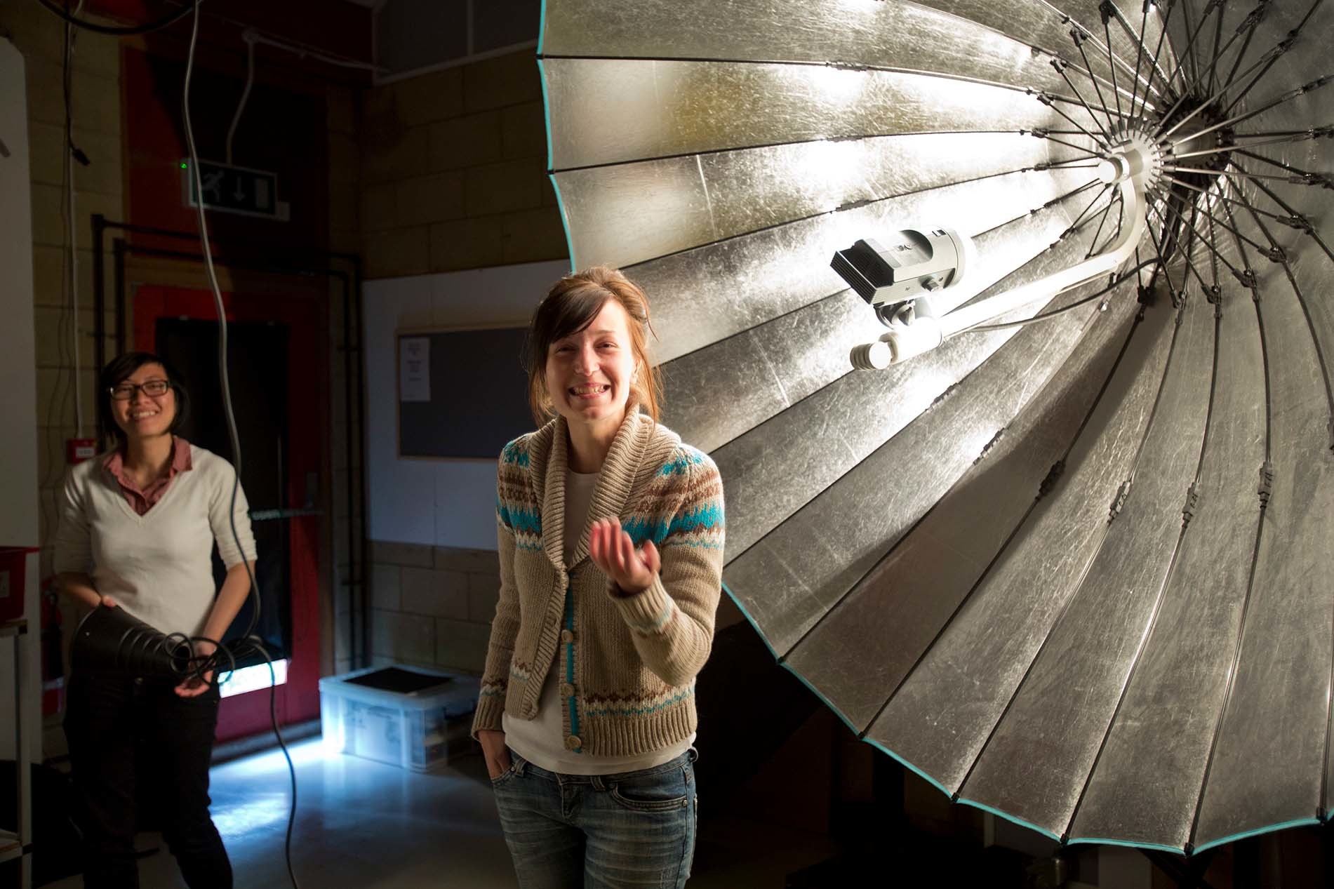 Two people stand smiling in a photo studio with a large piece of reflective photo equipment in the background to the right.