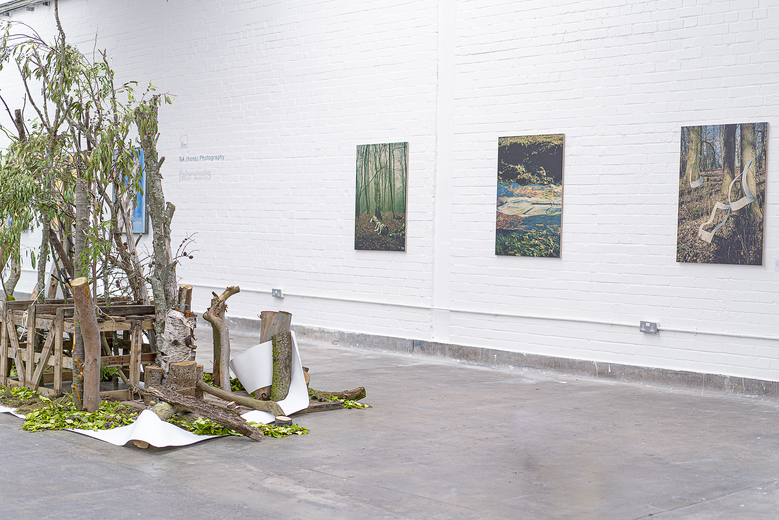 A sculpture made up of wooden pieces and trees sits in the middle of a gallery space. Three paintings hang on an exposed-brick wall.