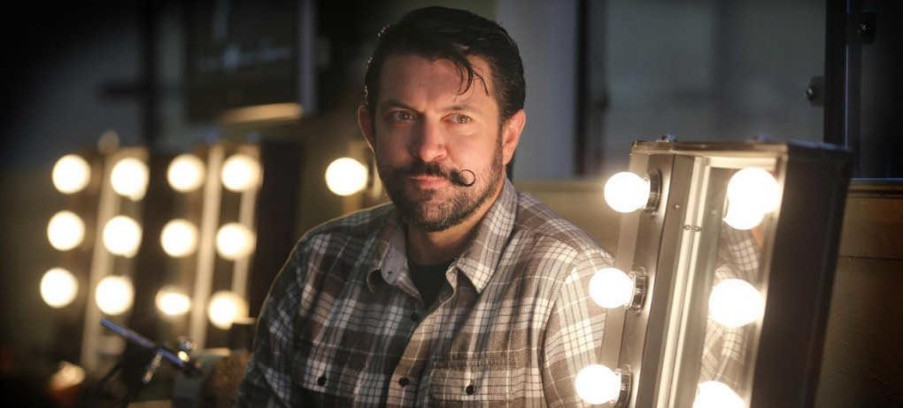 Headshot of B Kinney, sitting alongside a row of make-up mirrors with flashbulbs.