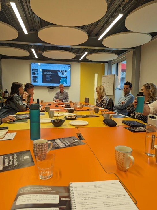 Business meeting in modern conference room with orange table, circular ceiling panels, wall-mounted screen, and people seated around table.