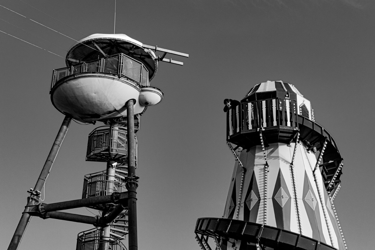 Two tall, futuristic amusement ride towers with enclosed passenger gondolas and spiral platforms against a clear sky.