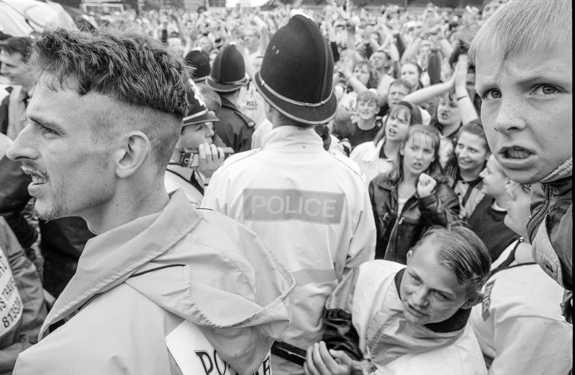 Black and white photo of a crowd at a football game, with uniformed police officers viewed from behind wading through the crowd.