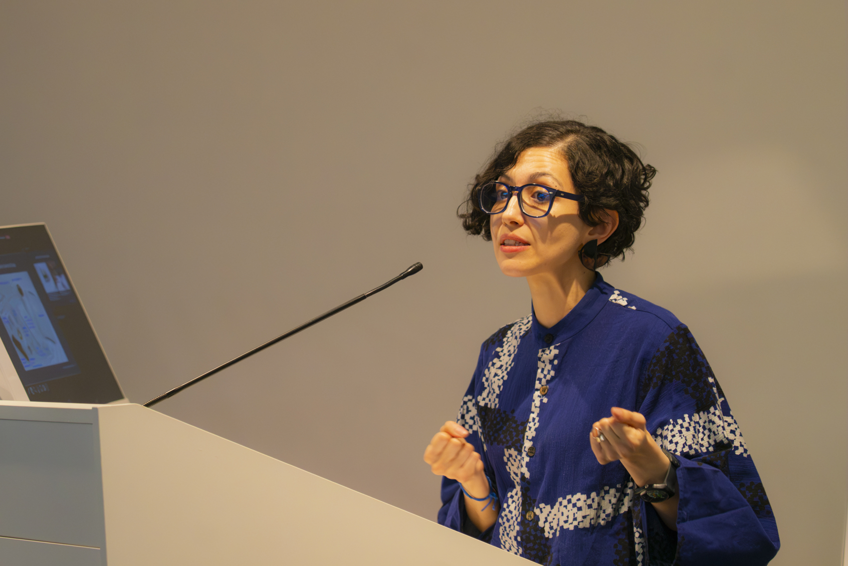 A person with curly black hair and glasses speaks at a podium, wearing a blue top with white patterns. In the background, a computer screen is visible, displaying information relevant to their presentation.