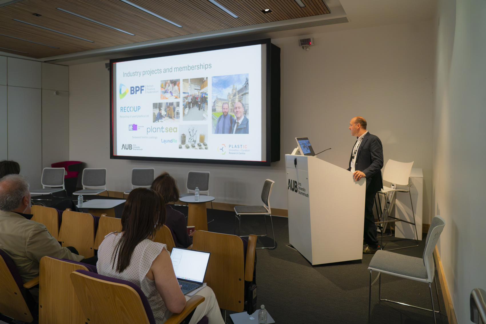A speaker stands at a podium presenting to an audience in a conference room. The screen behind shows logos for various industry projects and memberships. Attendees are seated, some using laptops, with wooden chairs and a bright interior.