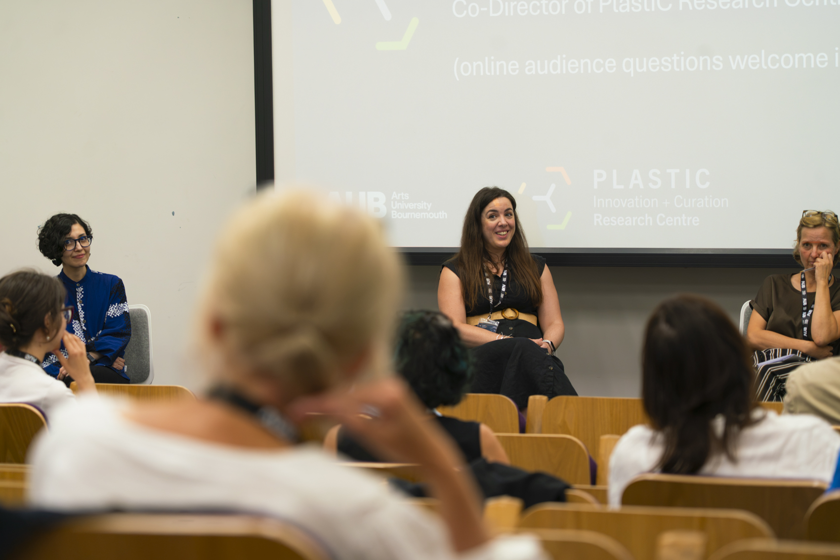A panel discussion taking place in a lecture hall, featuring three people seated behind a wooden stage. One woman, wearing a black top, speaks while the others listen, with an audience partially visible in the foreground.
