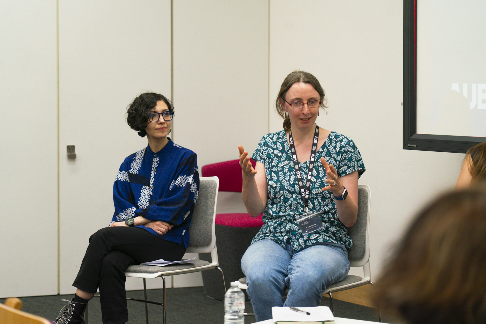 A person with glasses and curly hair in a blue patterned top sits on a chair, while another person in a green patterned top gestures with their hands while speaking. Both are in a modern room with a screen in the background and chairs in the foreground.