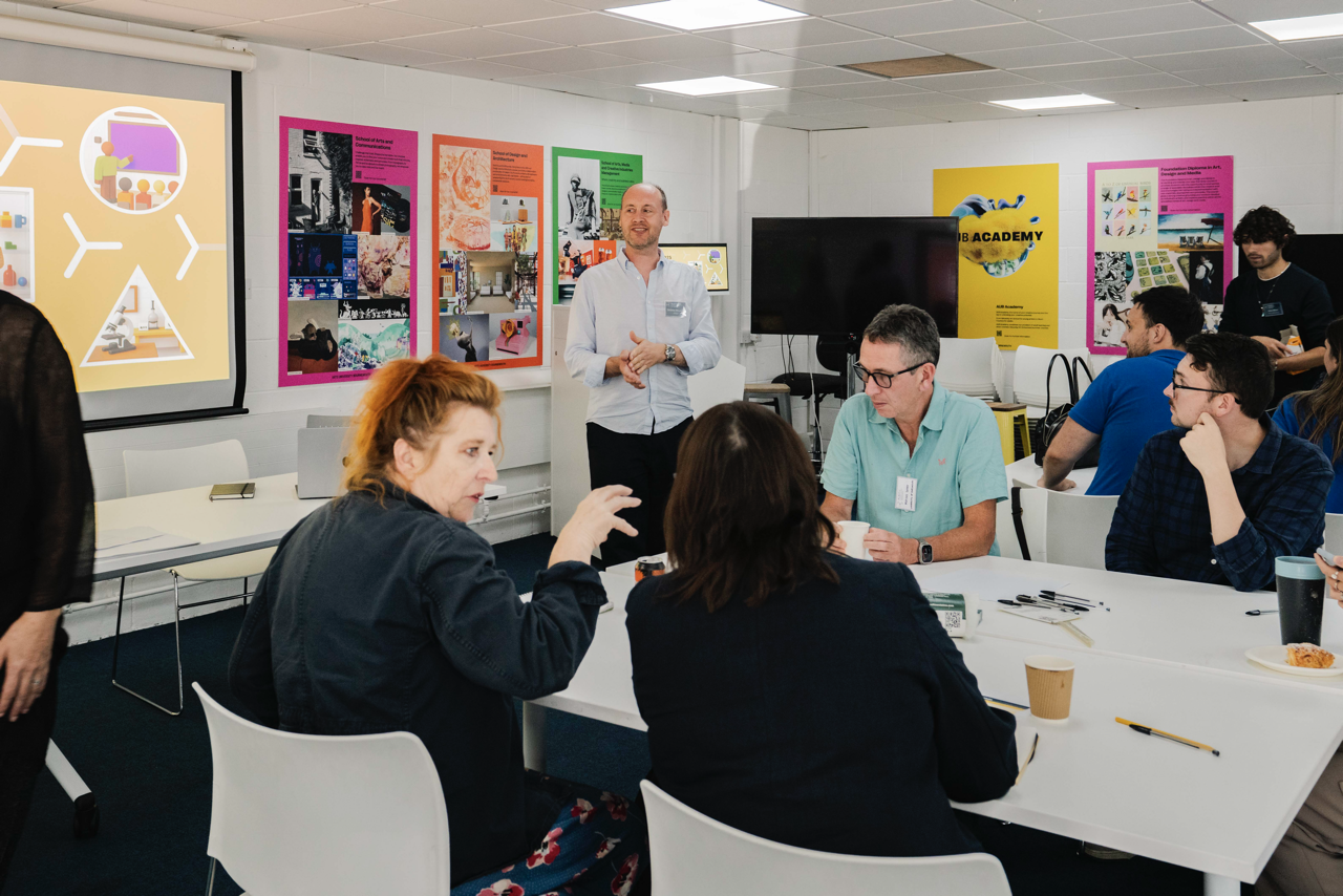 Group meeting in classroom with colourful posters on walls, people seated at white tables, one person standing presenting.