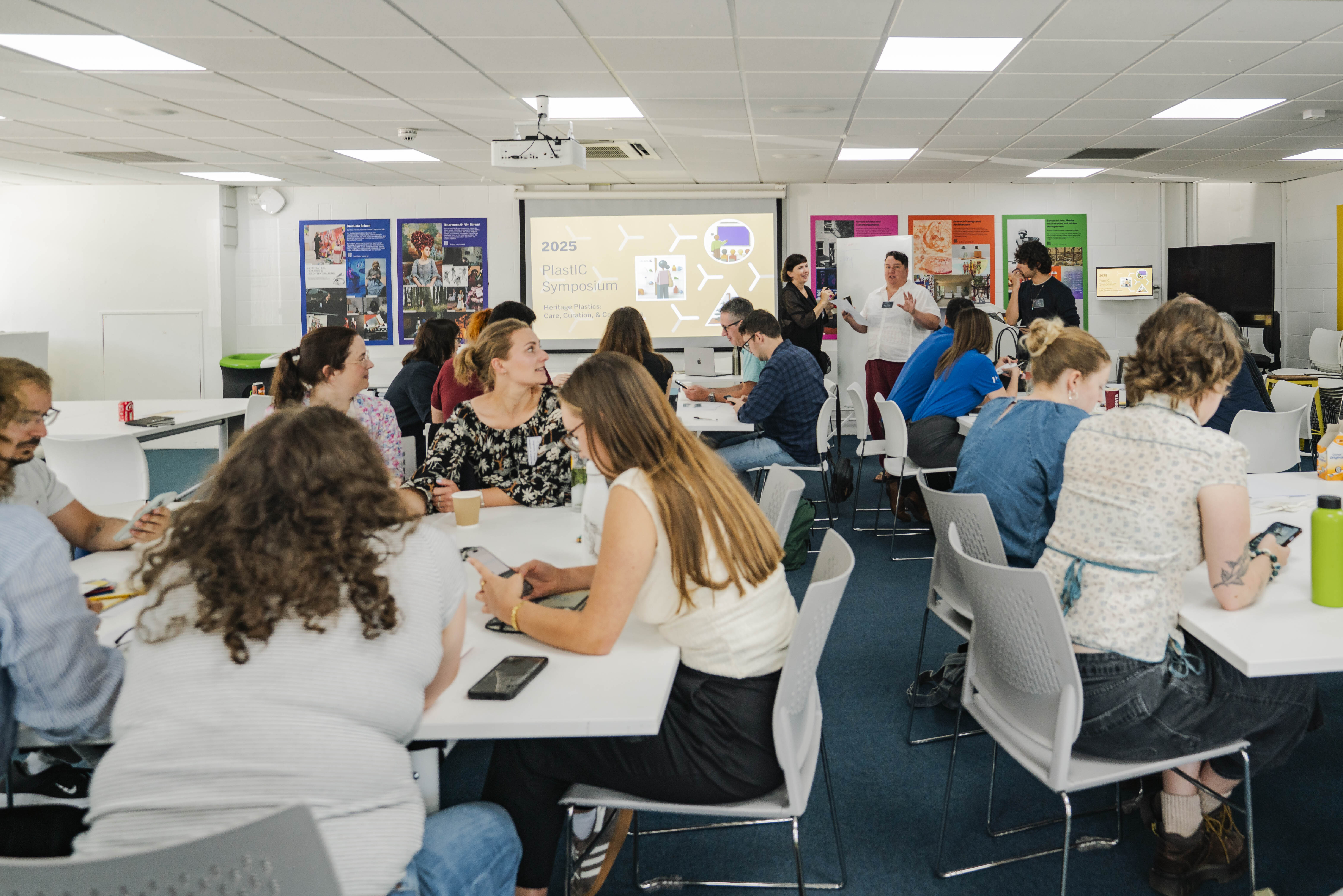 Students seated at white tables in modern classroom with fluorescent lighting, colourful wall displays, and teacher standing at front.