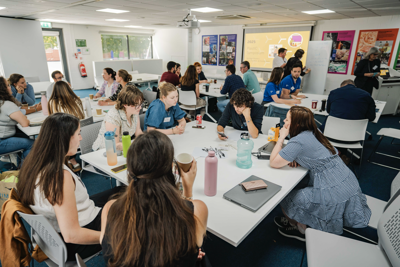 Students seated at white tables in classroom with fluorescent lighting, colourful wall displays, and windows along left wall.