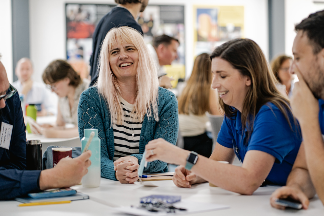 Blonde woman in striped top and teal cardigan sits at table with brunette woman in blue top, both smiling during group meeting.