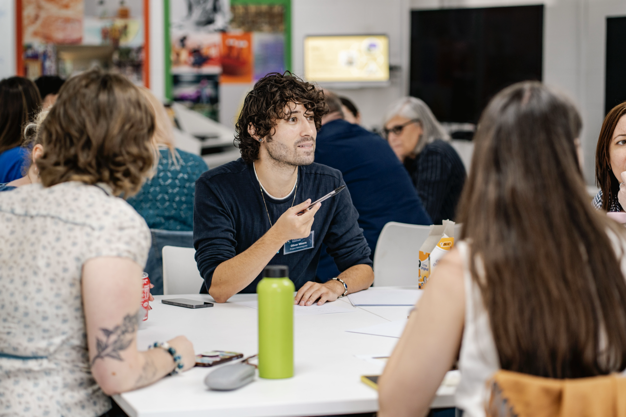 Young man with curly hair in dark jumper sits at white table with green water bottle, surrounded by people in classroom setting.