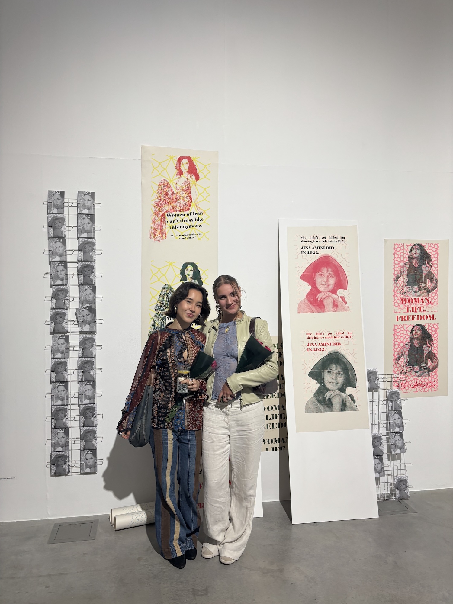 Two people posing together in front of white wall displaying colourful posters with portraits and black-and-white photo strips.