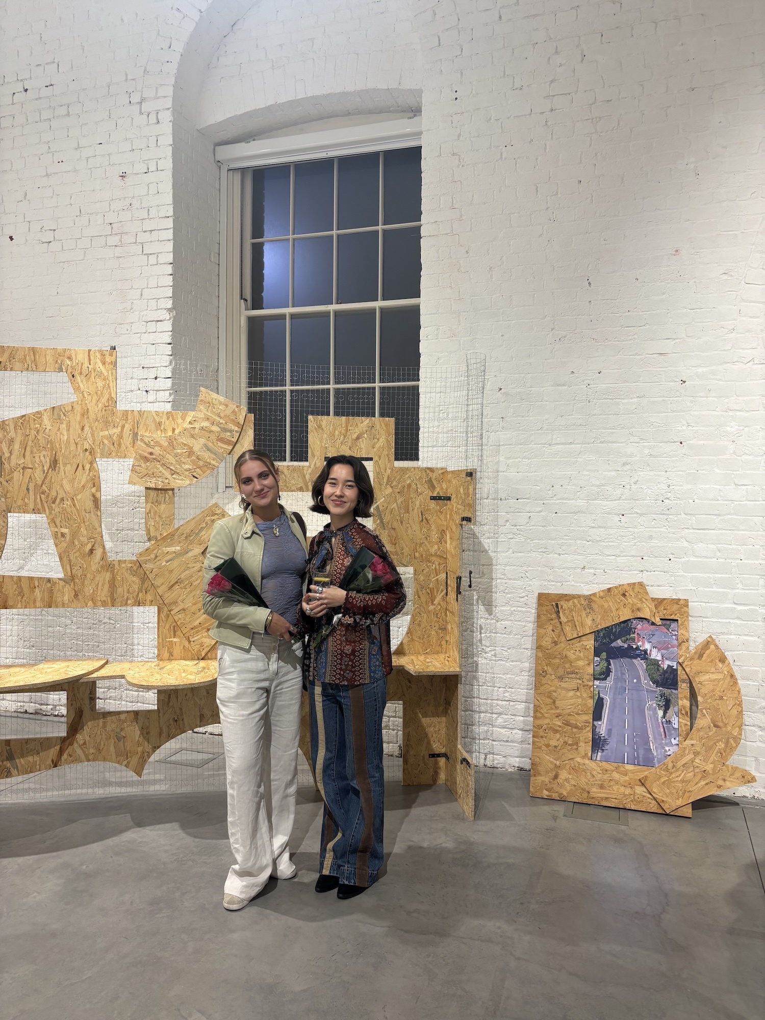 Two women standing in gallery space with yellow geometric sculptural installations against white brick wall with arched window.