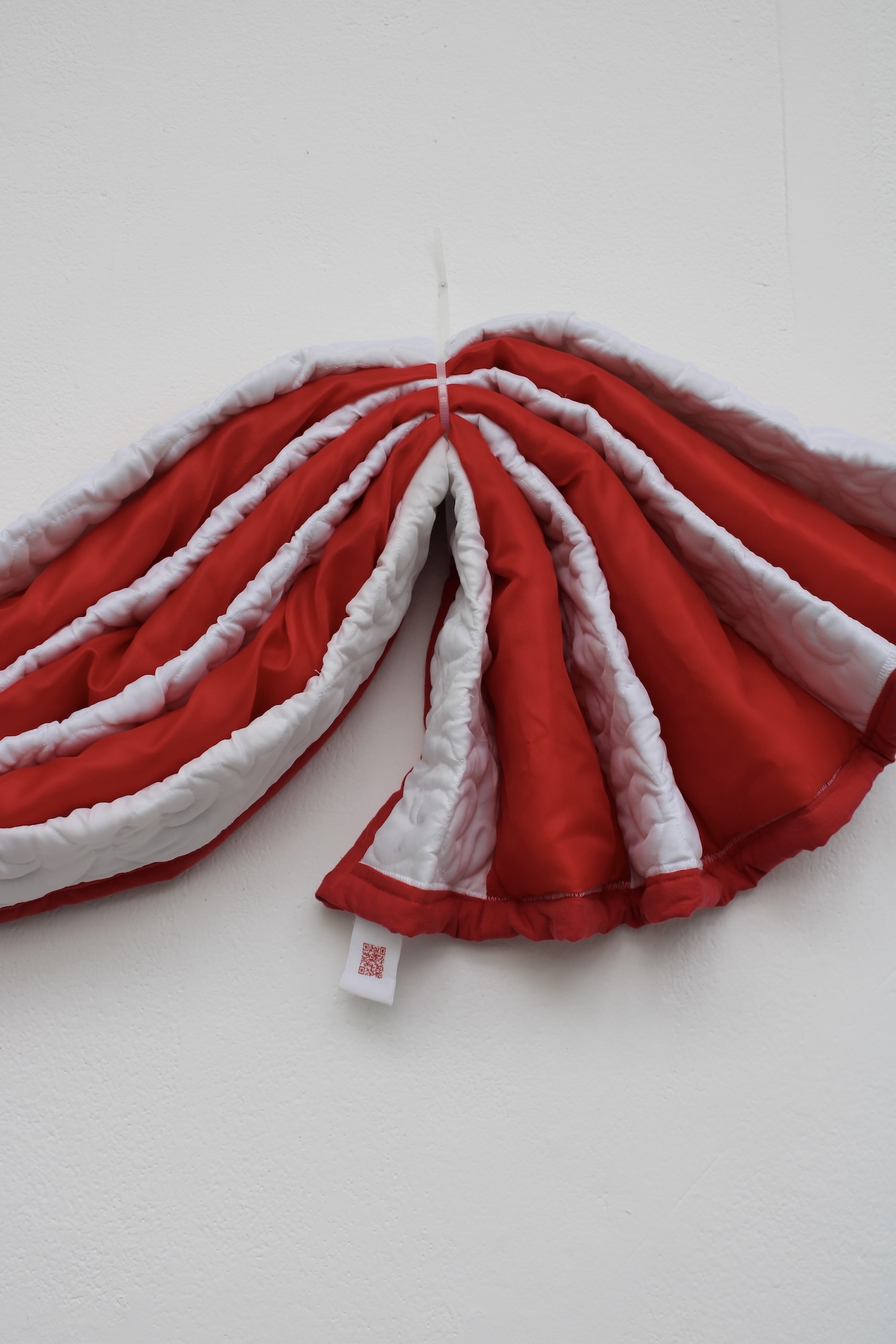 A red-and-white pleated fan-shaped garment or fabric piece hangs against a plain white wall, with a small tag visible at the bottom.