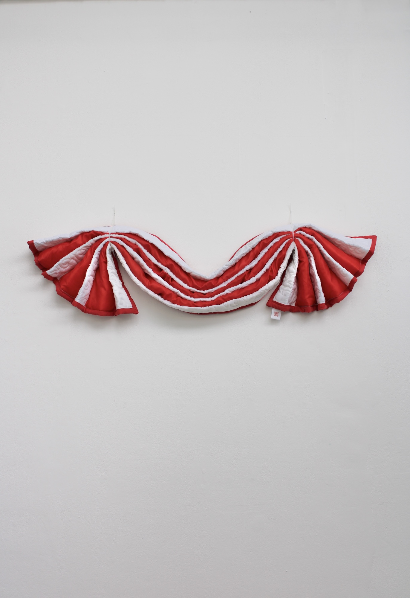 A red and white fabric garland with pleated folds hangs against a plain light grey wall.
