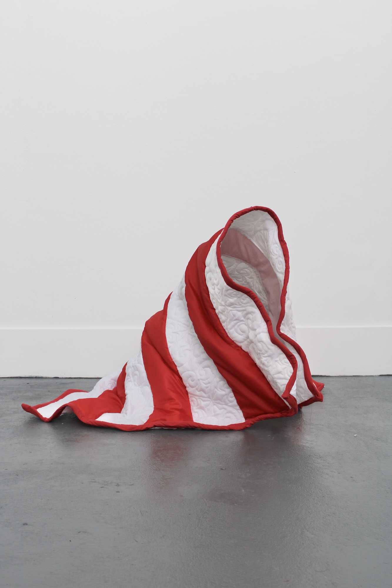 A red-and-white striped fabric sculpture draped on a grey floor against a plain white wall.