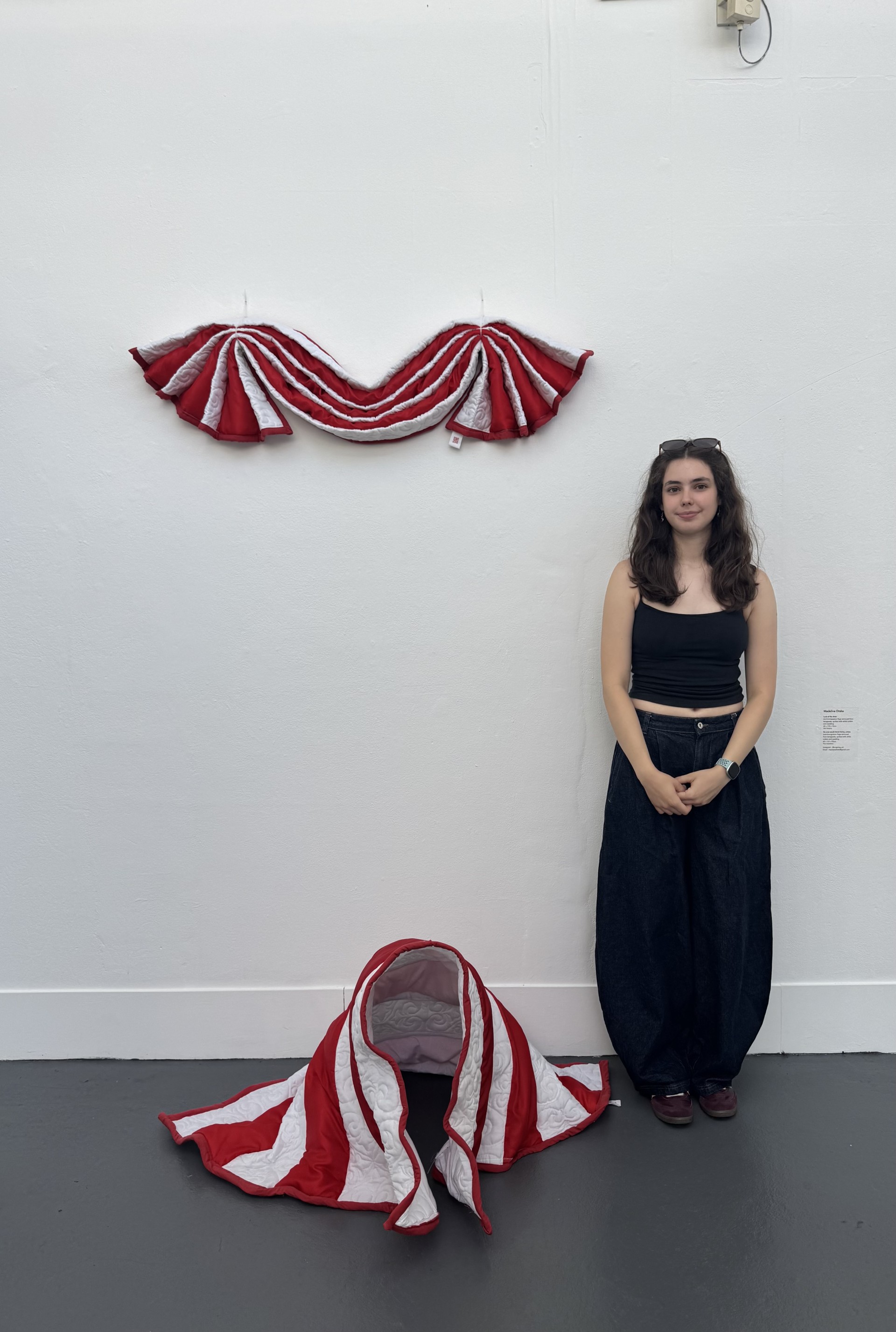 An Arts University Bournemouth graduate stands beside a red and white striped fabric sculpture, with a matching wall-mounted piece above it in a white gallery space.
