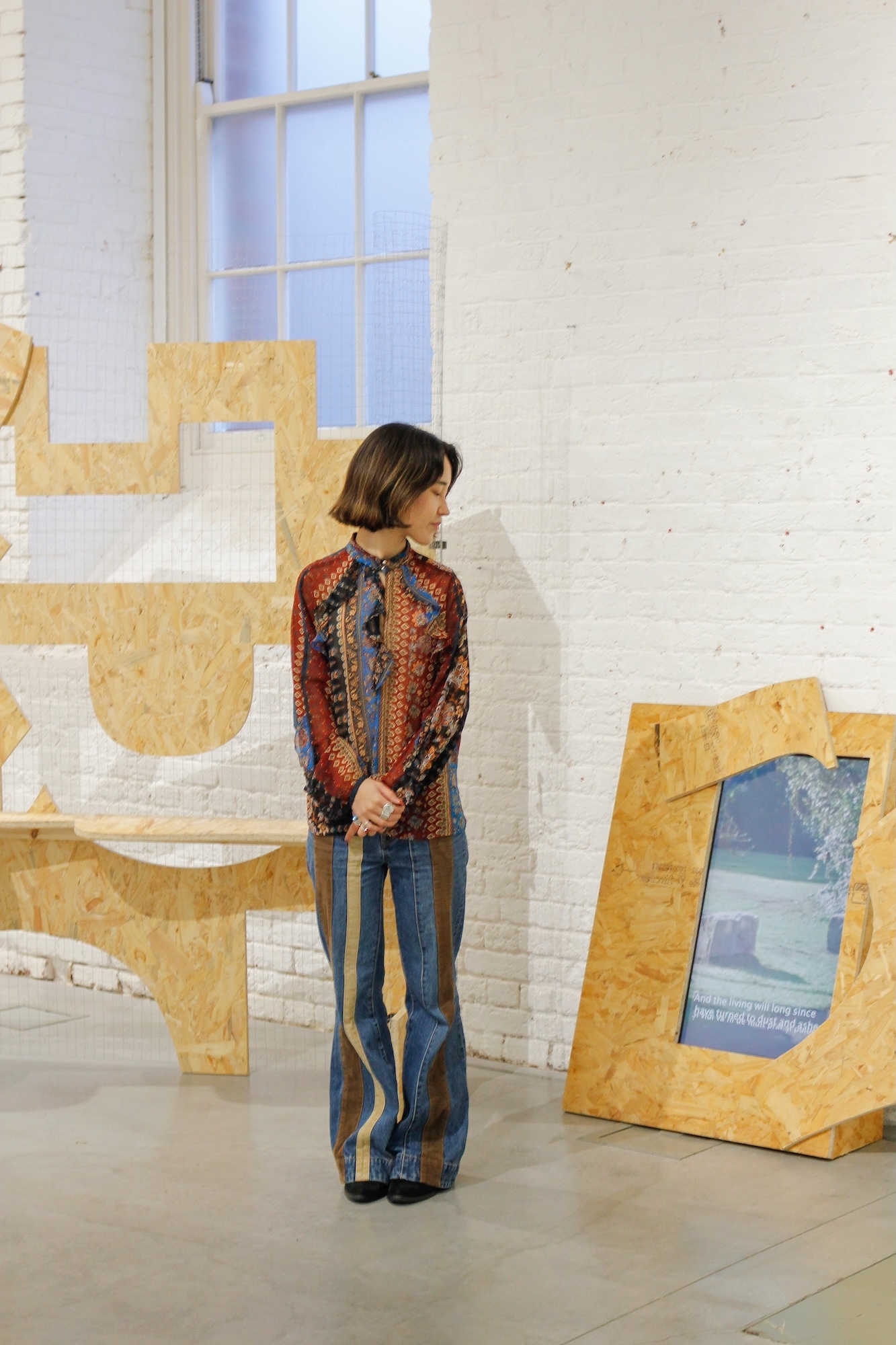 Woman in patterned shirt and flared jeans standing amongst large yellow geometric sculptures in white brick gallery space.