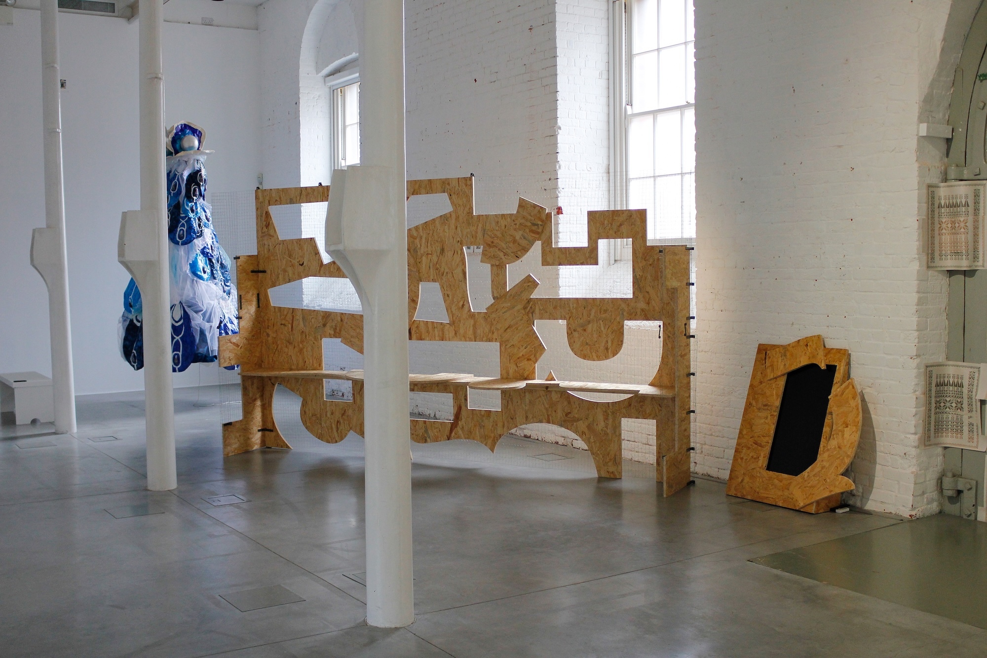 Gallery space with wooden geometric sculpture featuring interlocking angular forms, blue artwork on left wall, white columns and windows.