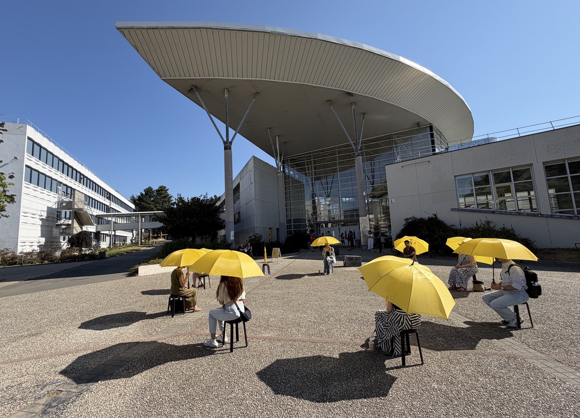 People sit on stools holding yellow umbrellas in front of a modern building with a large curved roof, under a clear blue sky.