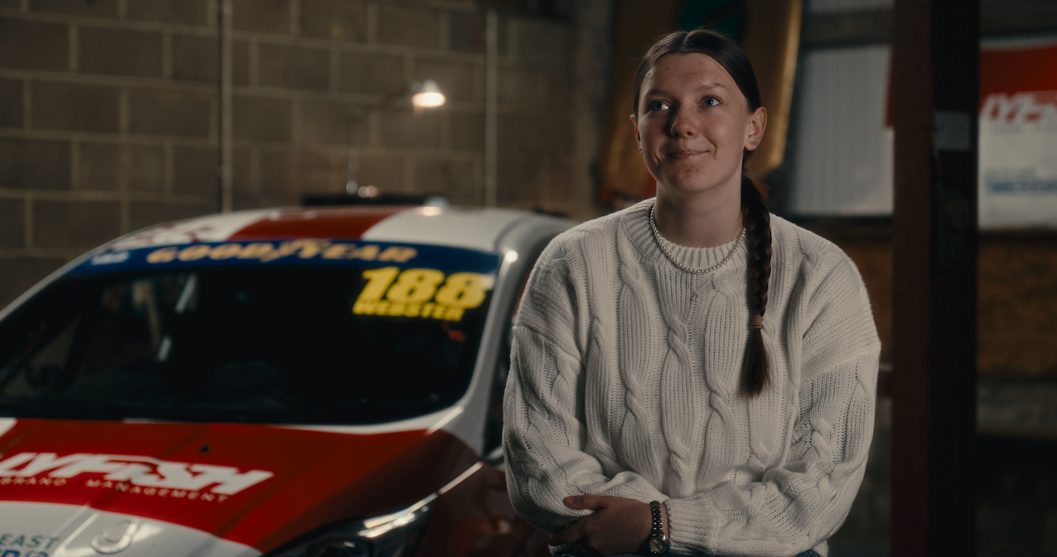 Woman in cream cable-knit jumper stands beside white racing car with red and blue stripes in garage setting.
