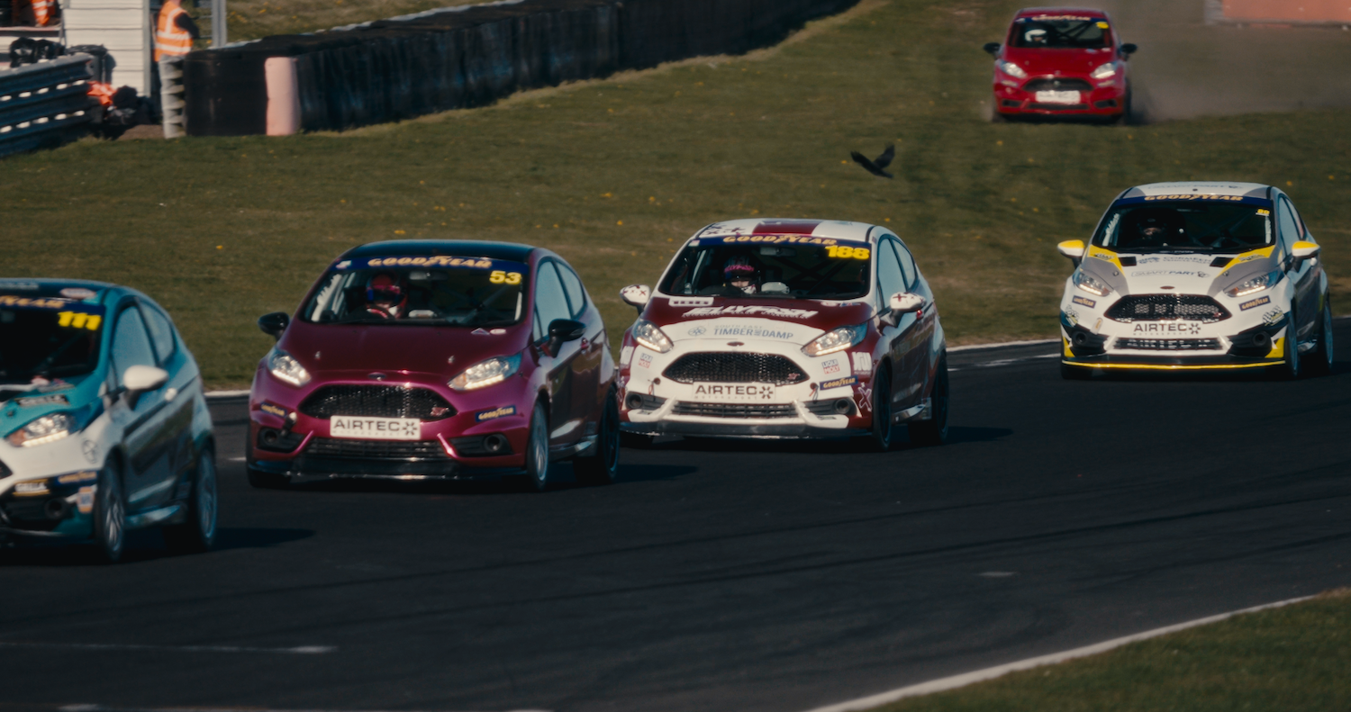 Racing cars competing on tarmac circuit, featuring red, white, and grey vehicles clustered together during race with green grass verge visible.