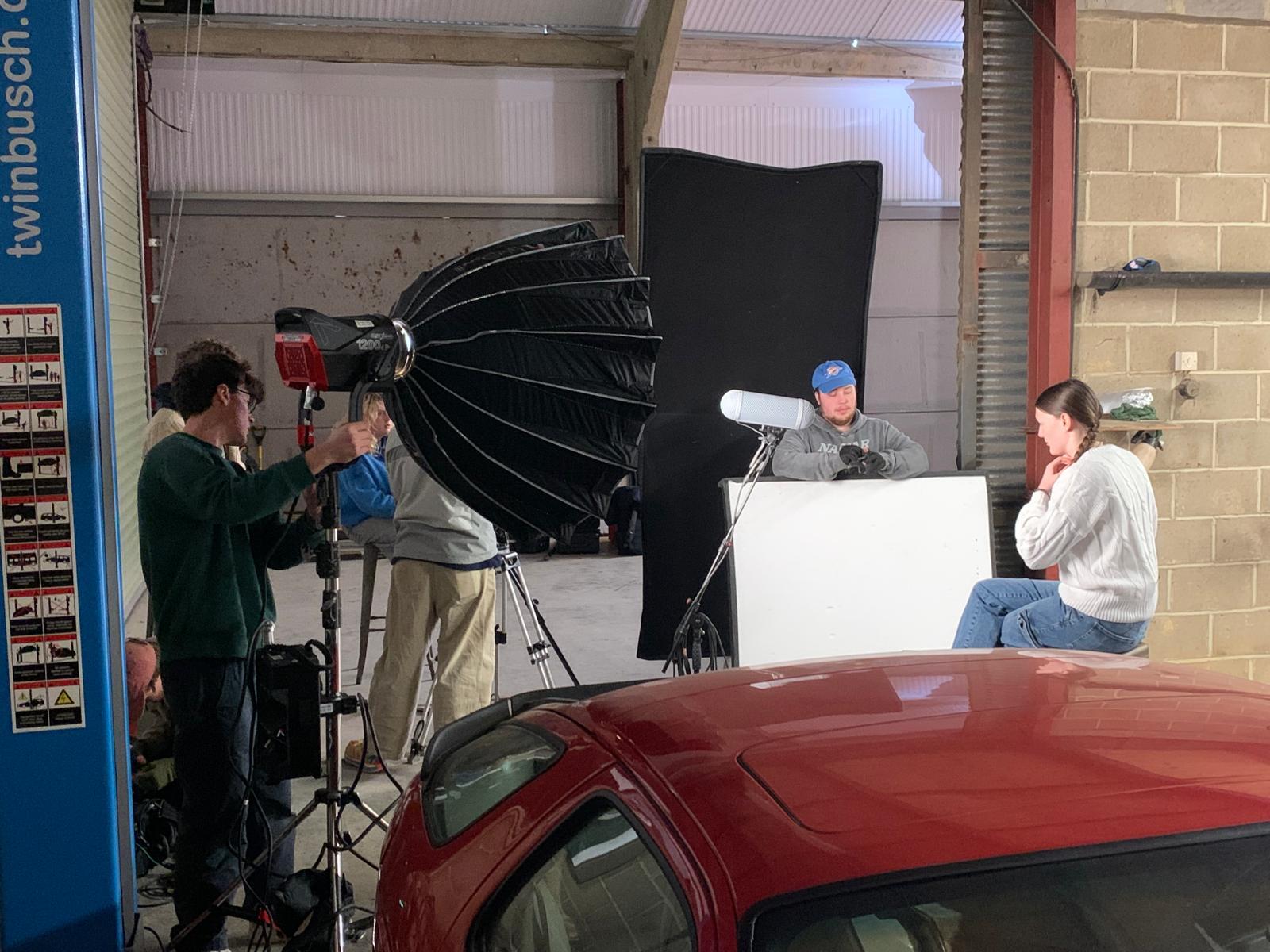 Film crew with cameras and large black umbrella light filming man at white desk in garage with red car in foreground.