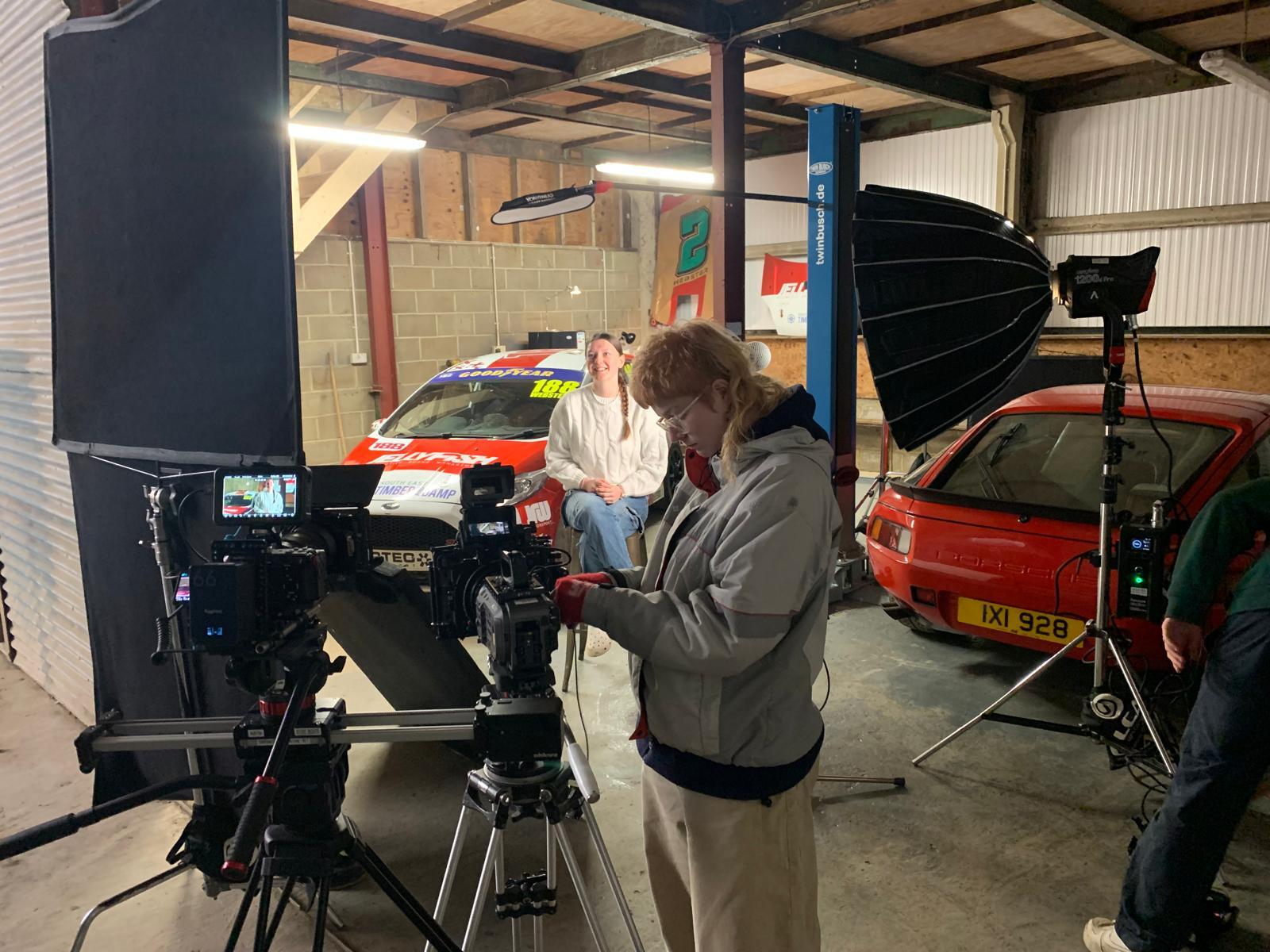 Cameraman operating professional camera equipment on tripod in garage workshop with classic cars and car lift in background.