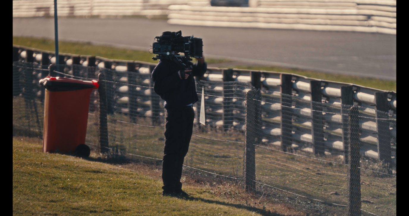Cameraman filming at race track beside tyre barrier wall, holding professional camera equipment on sunny day.
