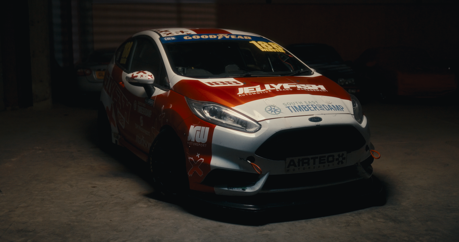 White rally car with red and blue sponsor decals parked in dimly lit garage or service area.