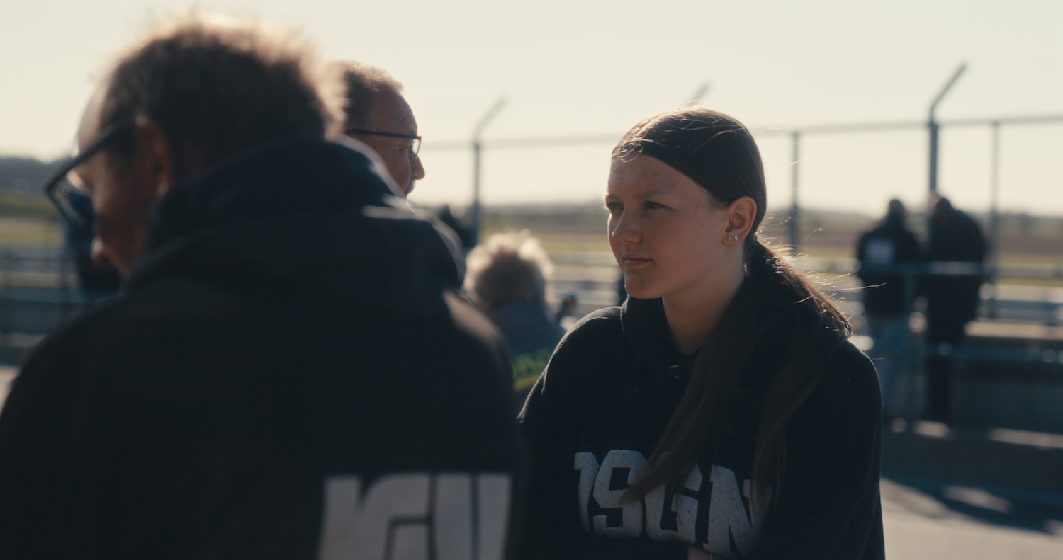 Woman in dark jacket speaking with person in foreground, industrial setting with cranes and structures in background, warm lighting.