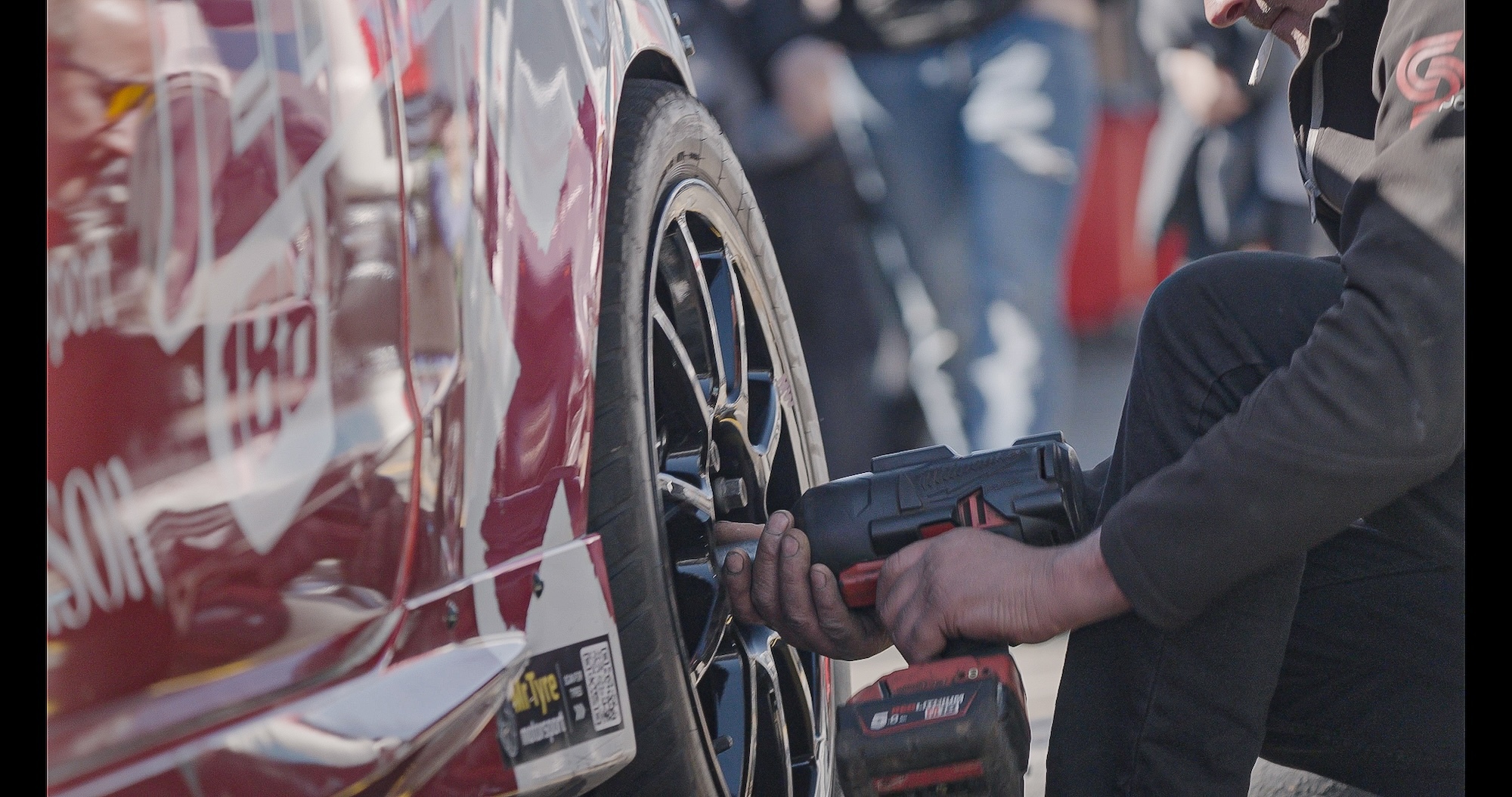 Person using pneumatic gun to change tyre on white race car with red graphics, crowd visible in blurred background.