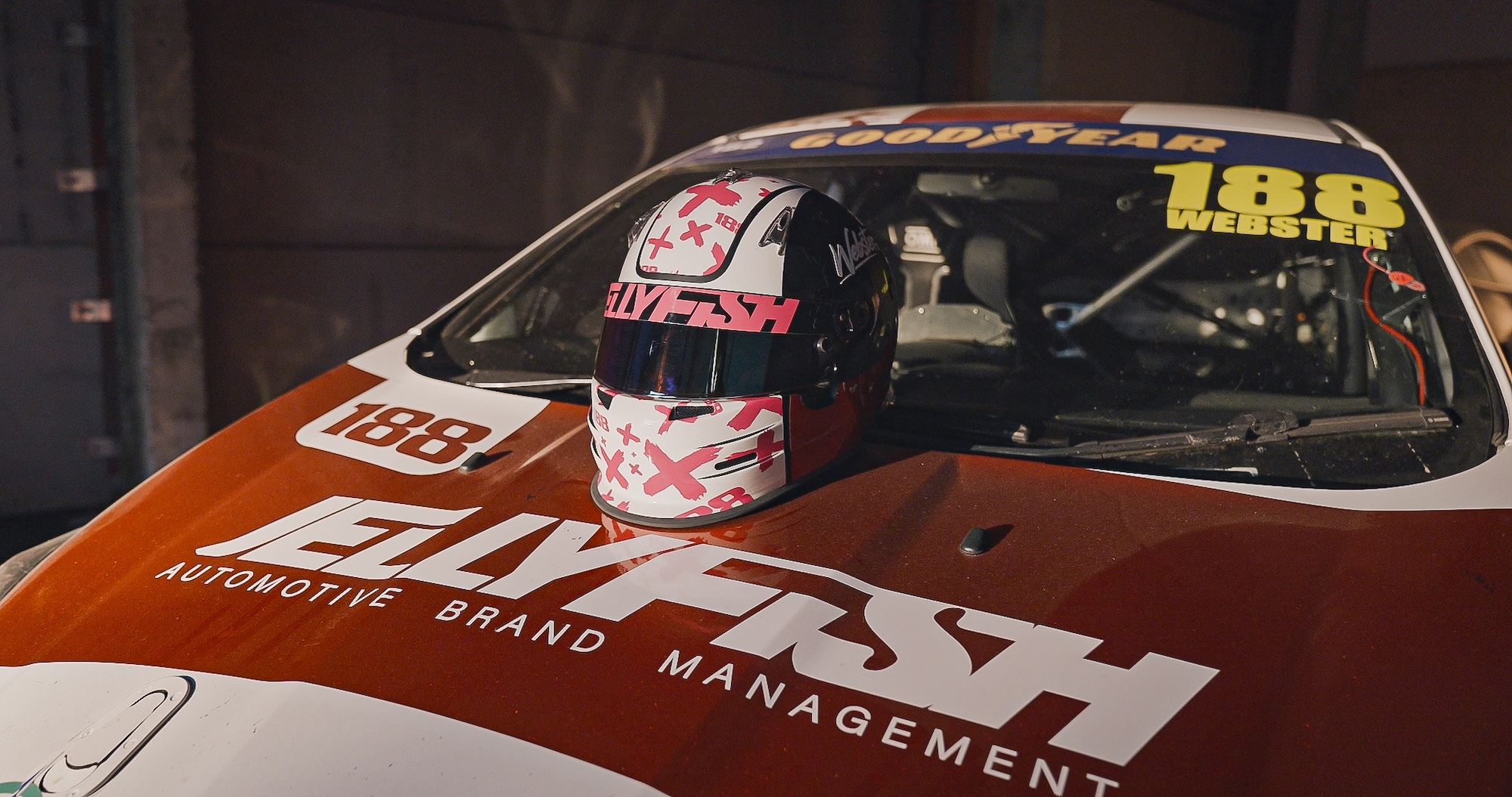Red and white racing car with number 188, driver wearing chequered helmet visible through windscreen, garage setting.