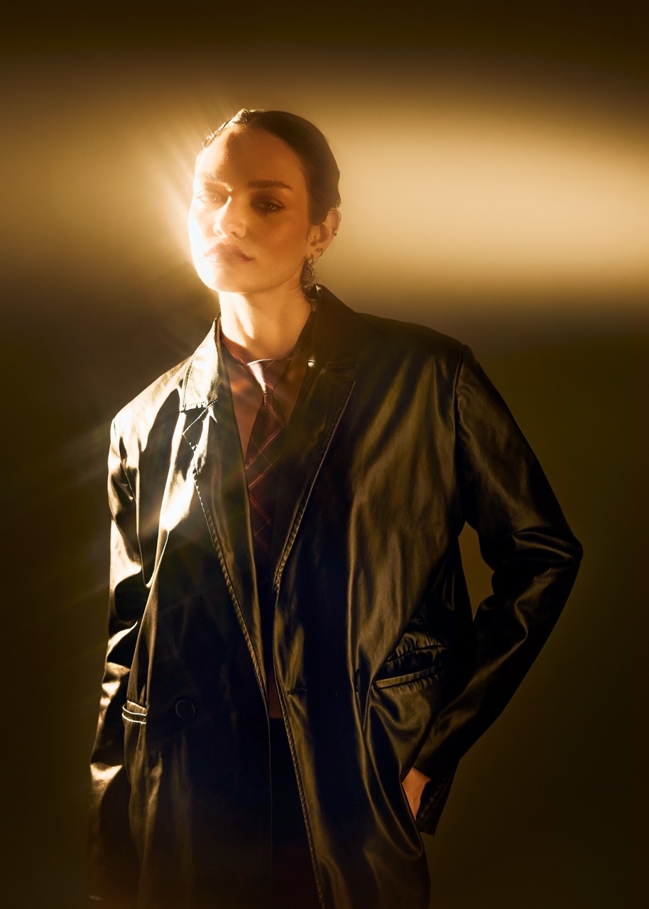 A woman wearing a black leather blazer stands against a dark background, illuminated by a beam of light emanating from behind her. The light highlights her profile and creates a soft glow around her shoulder.