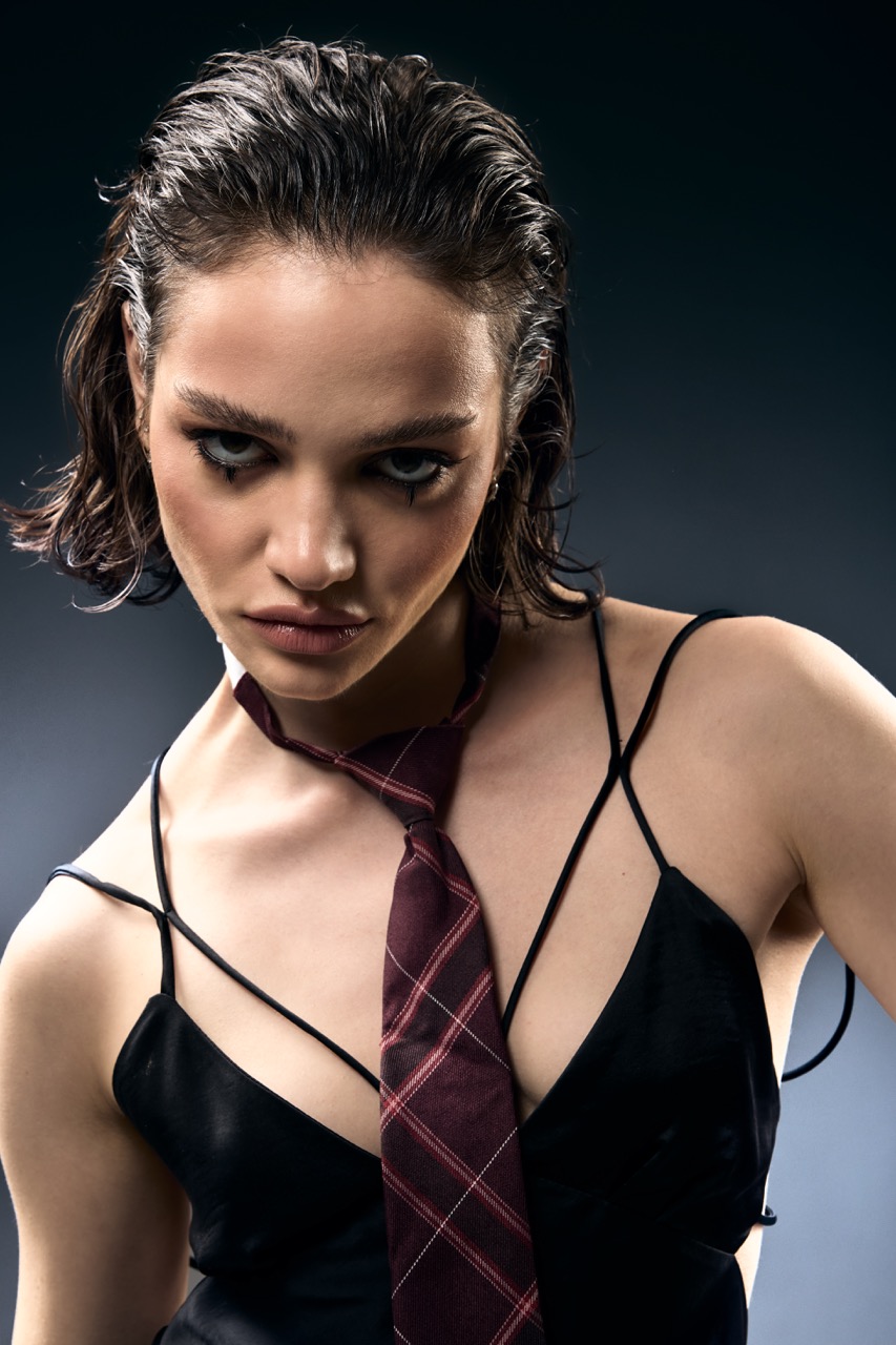 A young woman with wet hair is wearing a black sleeveless top and a maroon tie. She is positioned slightly to the side, with her head tilted down, and the background features a gradient from dark to light.