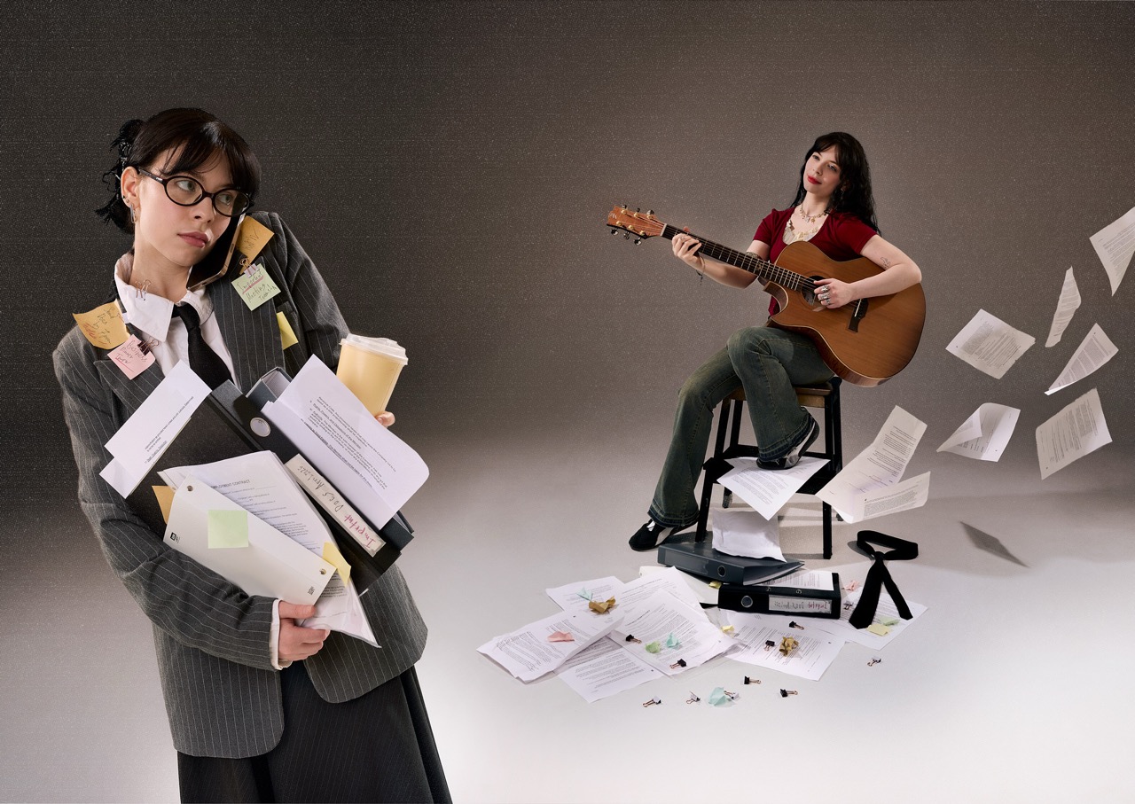 A woman in a grey blazer, holding stacks of papers and a coffee cup, stands on the left. On the right, another woman sits on a stool playing an acoustic guitar, surrounded by scattered sheets of paper on the floor.
