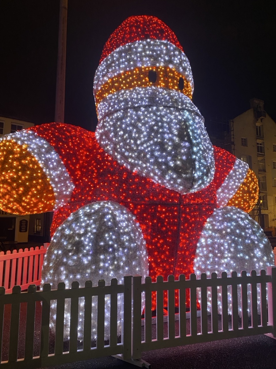 Large illuminated elephant sculpture covered in red, white and orange LED lights, surrounded by white picket fence at night.