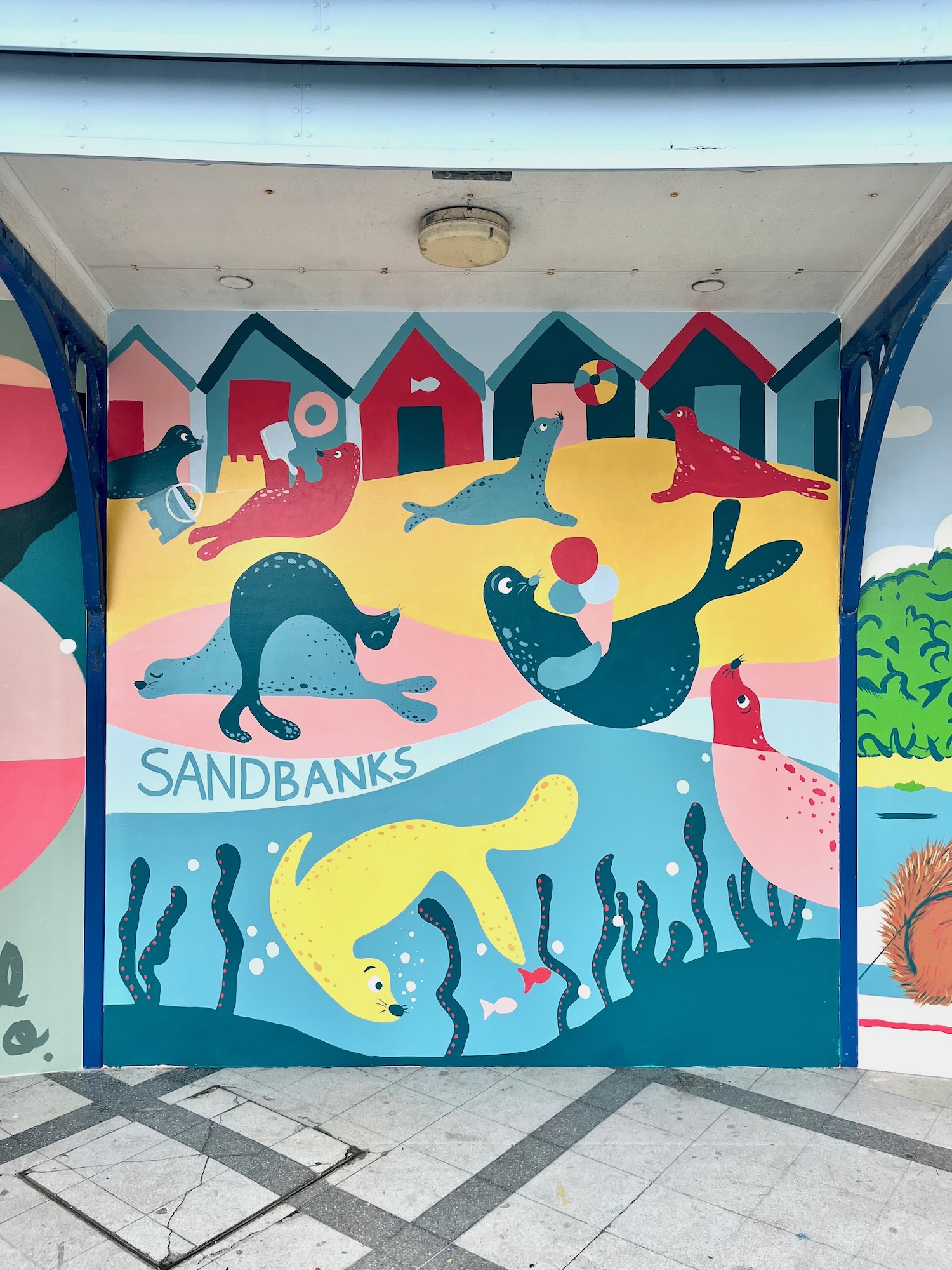A colourful mural of sea lions and fish on a wall at Sandbanks, with stylised beach huts and seaweed above a tiled floor.