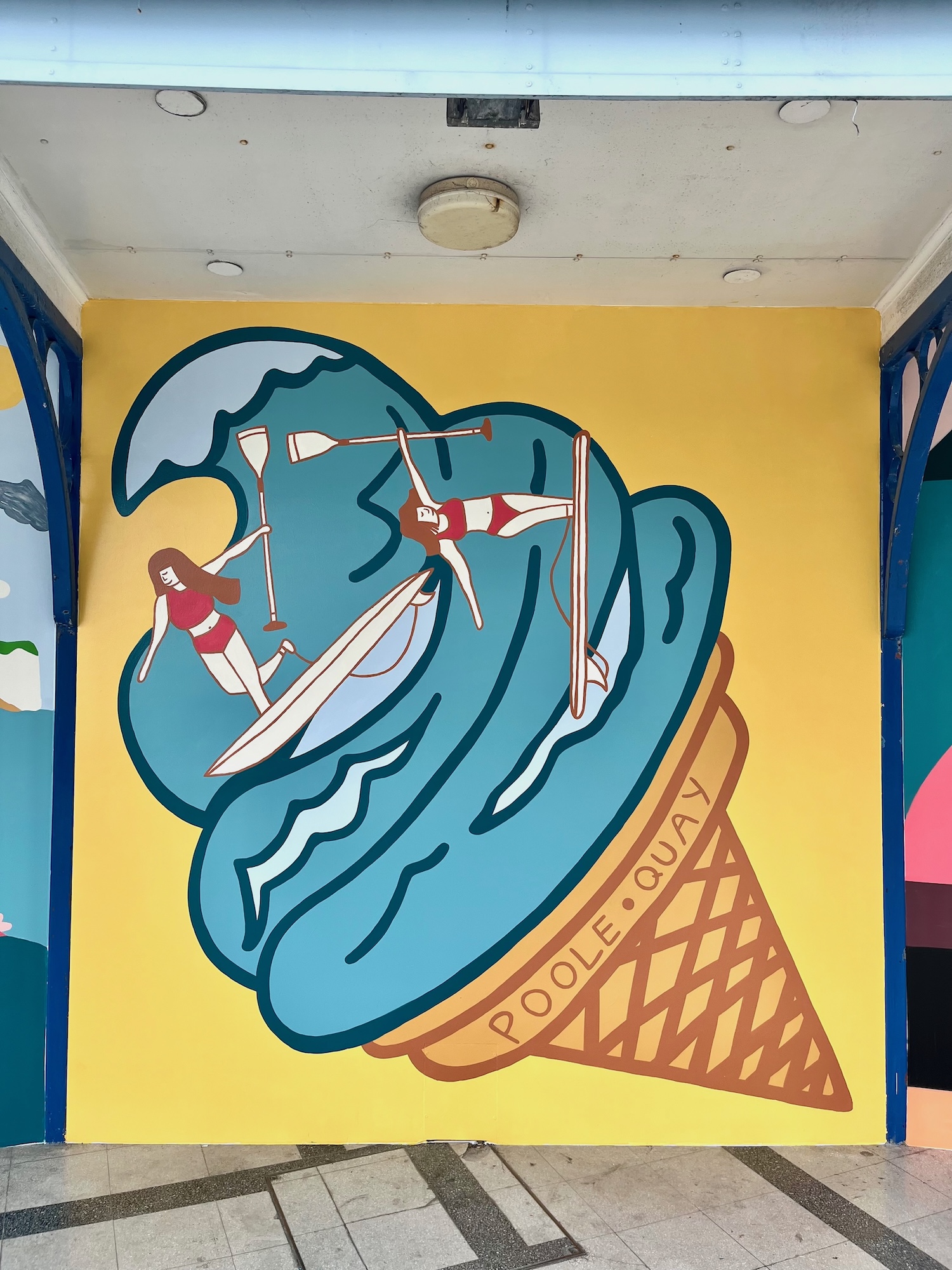 A colourful mural of two paddleboarders riding a large blue wave painted on a yellow wall, with a ‘POOLE’ ice-cream cone design below.